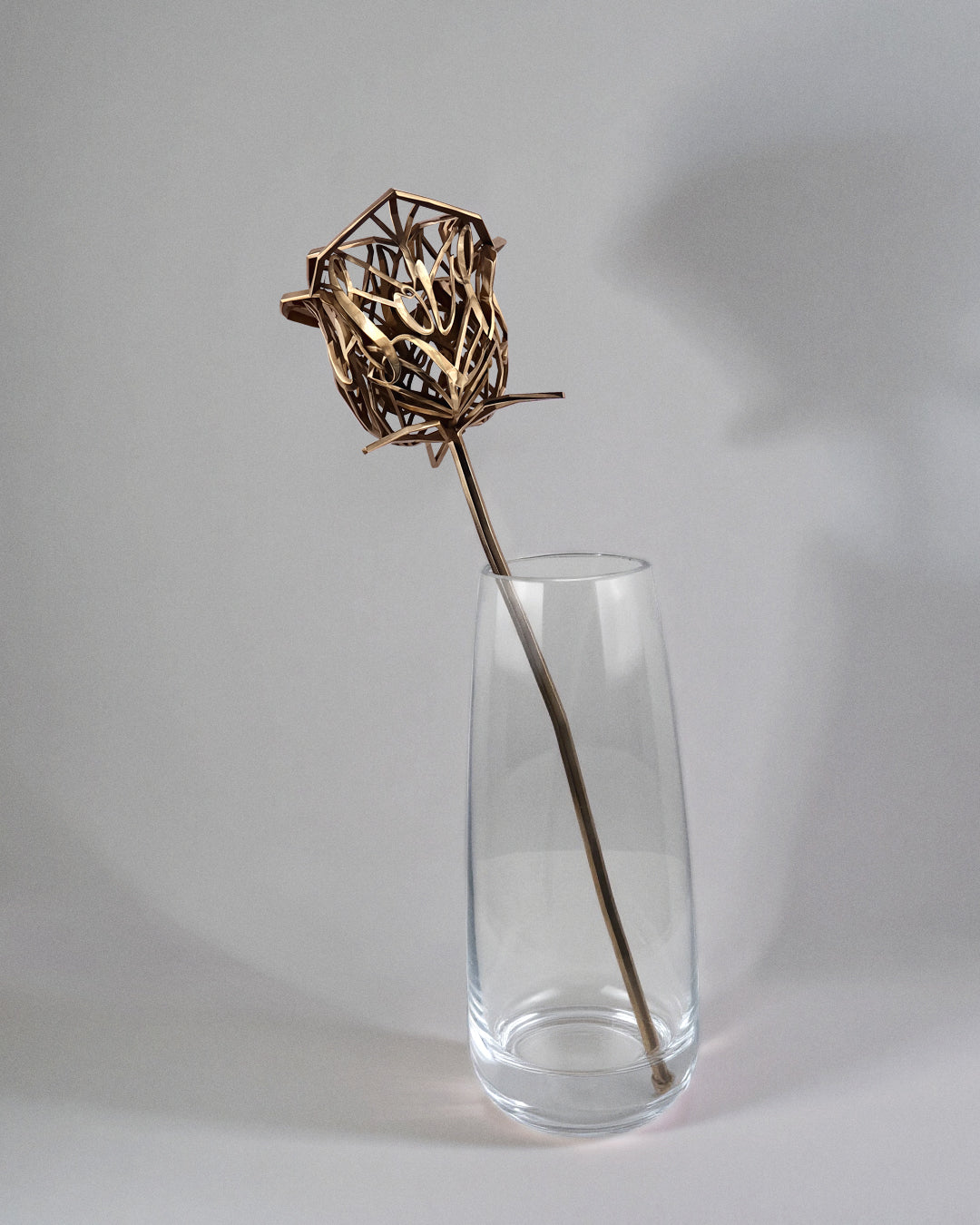 Bronze rose sculpture with letter-shaped petals displayed in a clear glass vase against a neutral background