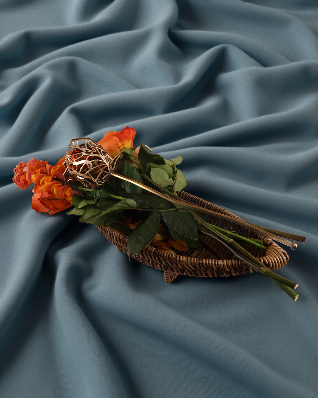 Bronze rose sculpture resting atop French blue drapery, paired with orange roses — a study in craftsmanship, contrast, and elegance